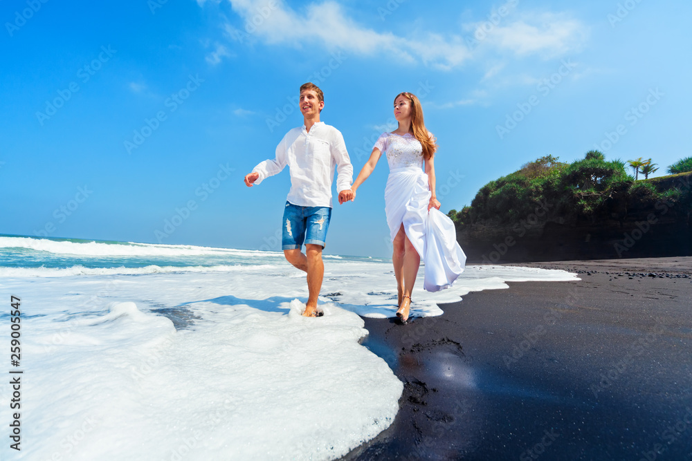 Happy family on honeymoon holiday - just married young man and woman run with fun by black sand beac