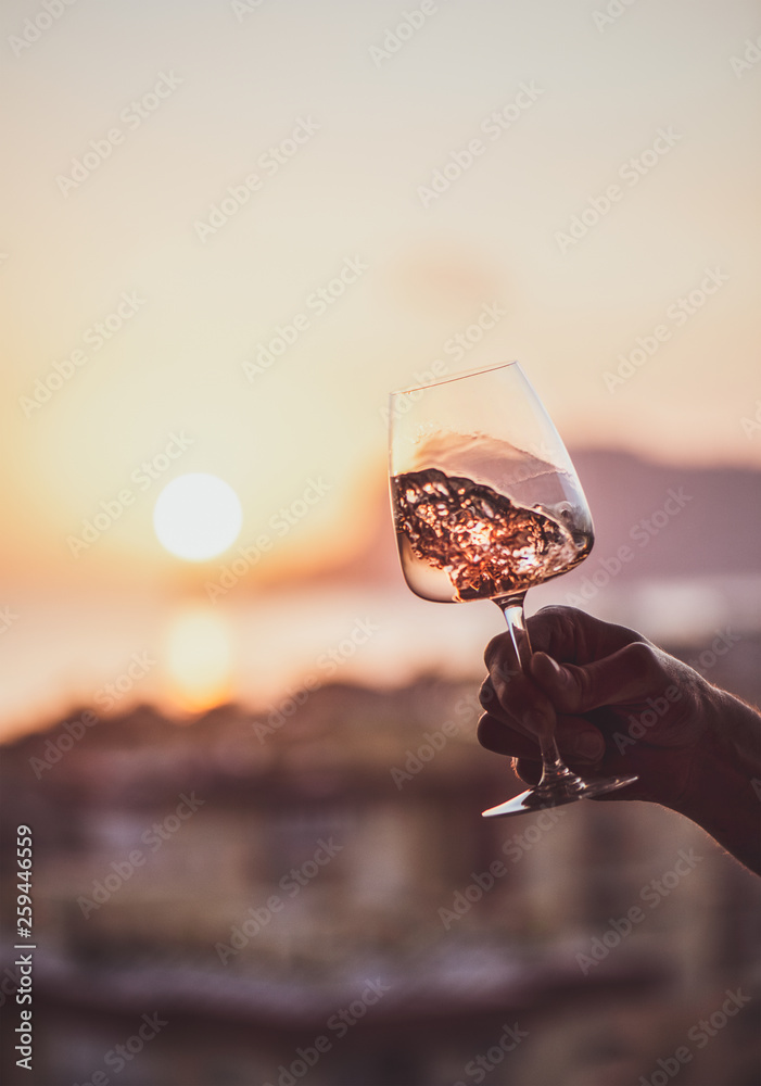 Mans hand holding glass of rose wine and with sea and beautiful sunset at background, close-up. Sum