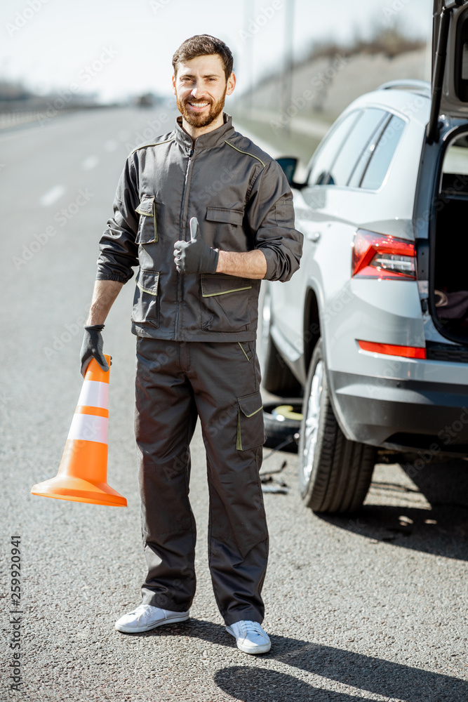 Portrait of a happy road assistance worker in uniform standing with cone near the car on the roadsid