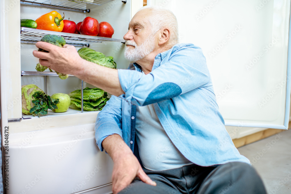 Handsome senior man taking fresh cabbage sitting near the refrigerator full of healthy products at h
