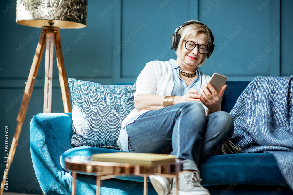 资深女士穿着休闲，在家的沙发上戴着耳机和手机听音乐