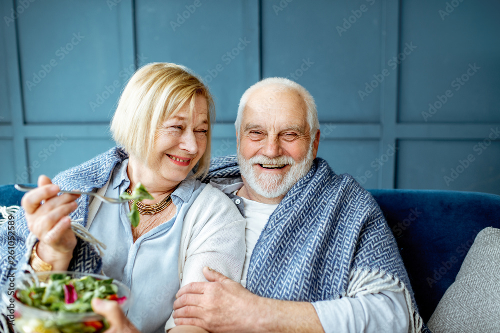 可爱的老年夫妇裹着格子布坐在家里的沙发上吃着健康的沙拉