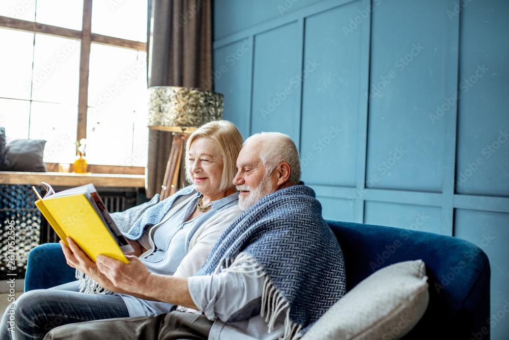 Lovely senior couple reading book while sitting wrapped with warm plaid on the couch at home