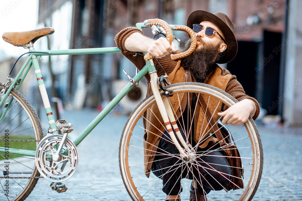一位留着胡子的潮人的肖像，他穿着时髦，戴着帽子，在户外的u形公路上拥抱着他的复古自行车