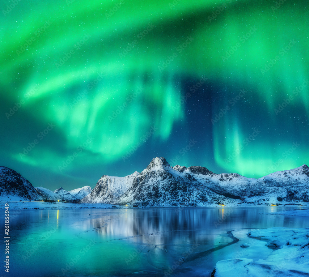 Aurora borealis over snowy mountains, frozen sea coast and reflection in water in Lofoten islands, N