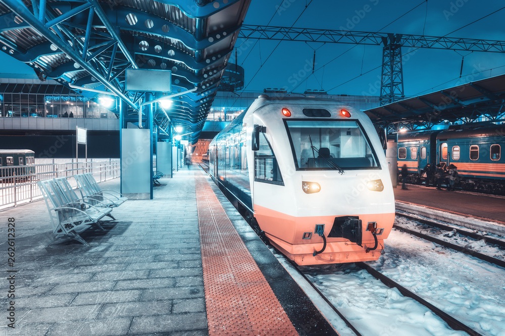 High speed train on the railway station at night in winter. Urban landscape with modern commuter whi