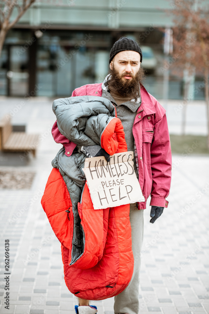 一个沮丧无家可归的乞丐拿着睡袋和纸板站在公共汽车旁的画像