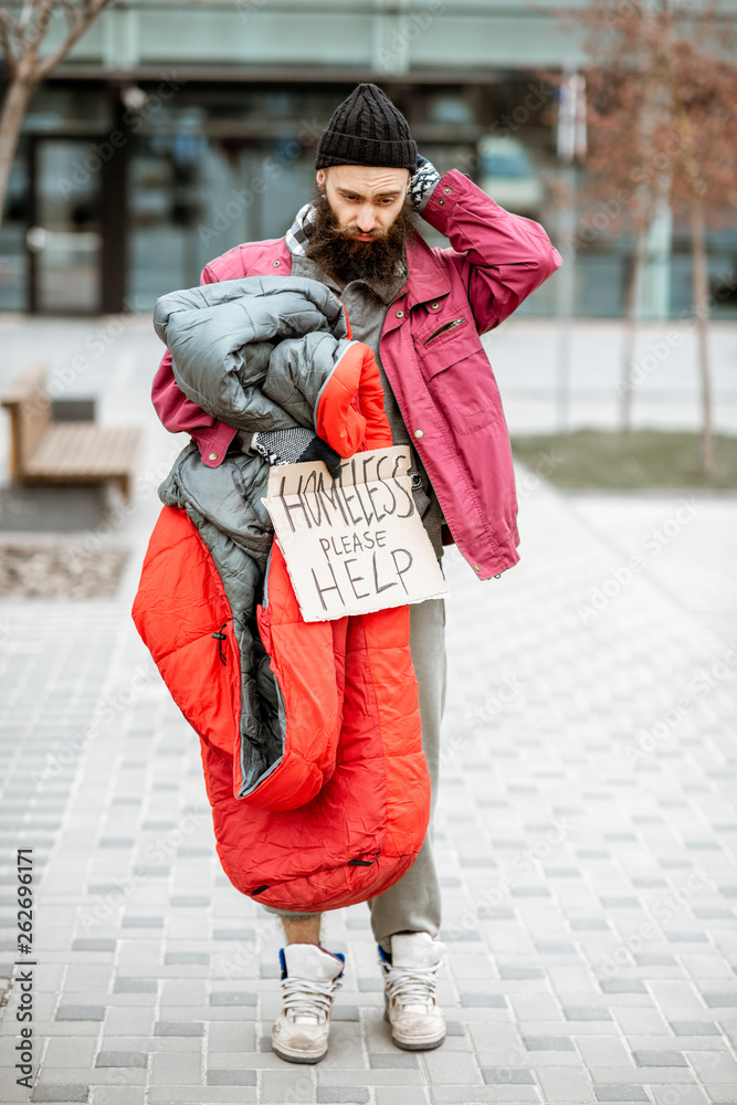 一个沮丧无家可归的乞丐拿着睡袋和纸板站在公共汽车旁的画像