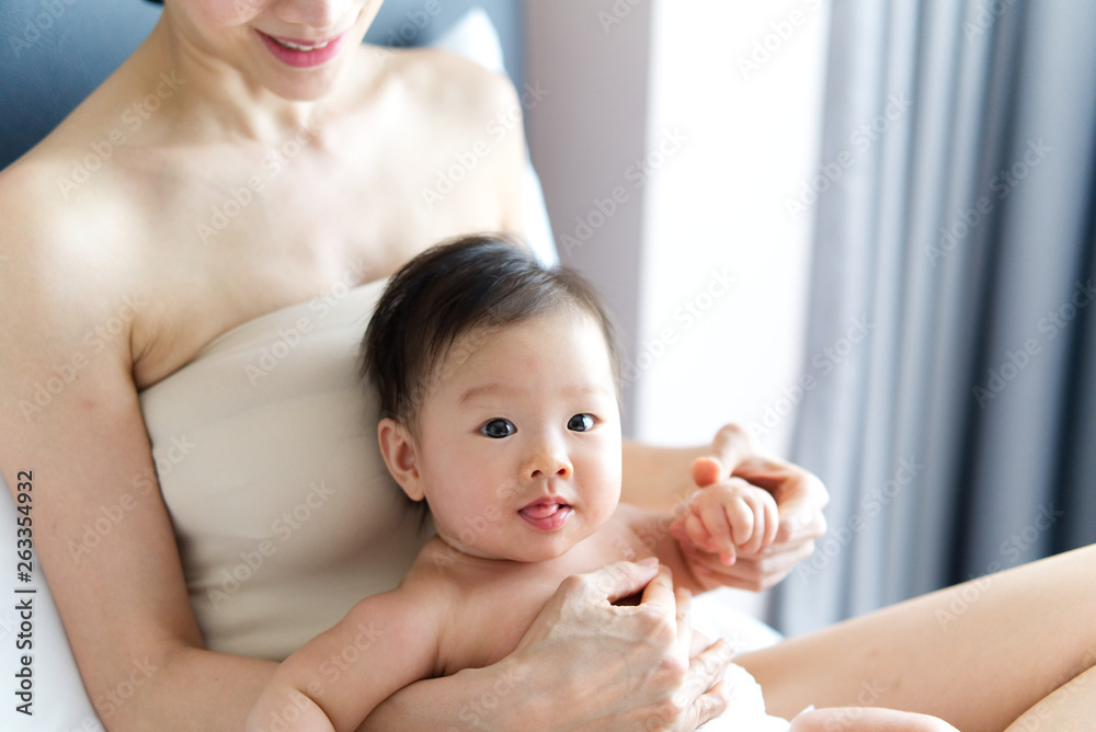 亚洲可爱的婴儿坐在床上母亲的腿上。婴儿在运动时看着镜头