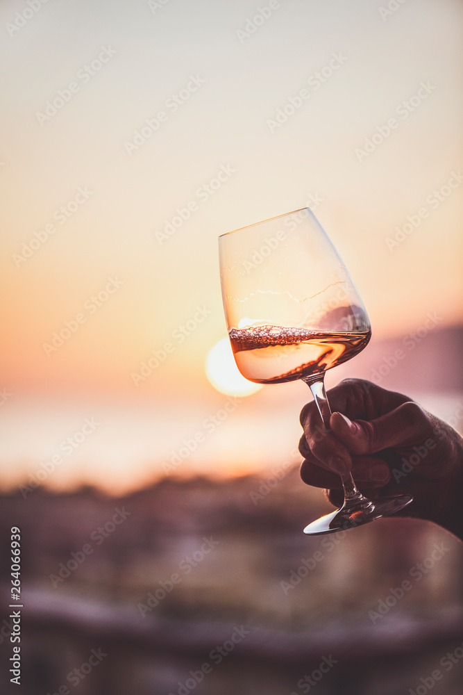 Glass of rose wine in mans hand with sea view and sunset at background, close-up. Summer evening 