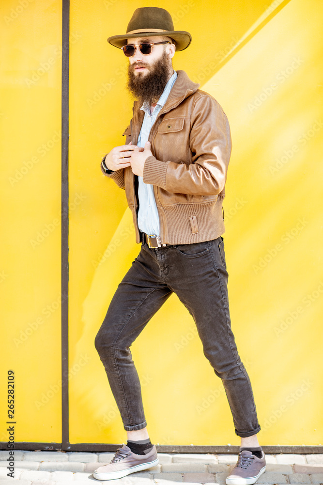 Full length portrait of a stylish bearded man dressed in jacket and hat on the bright yellow backgro