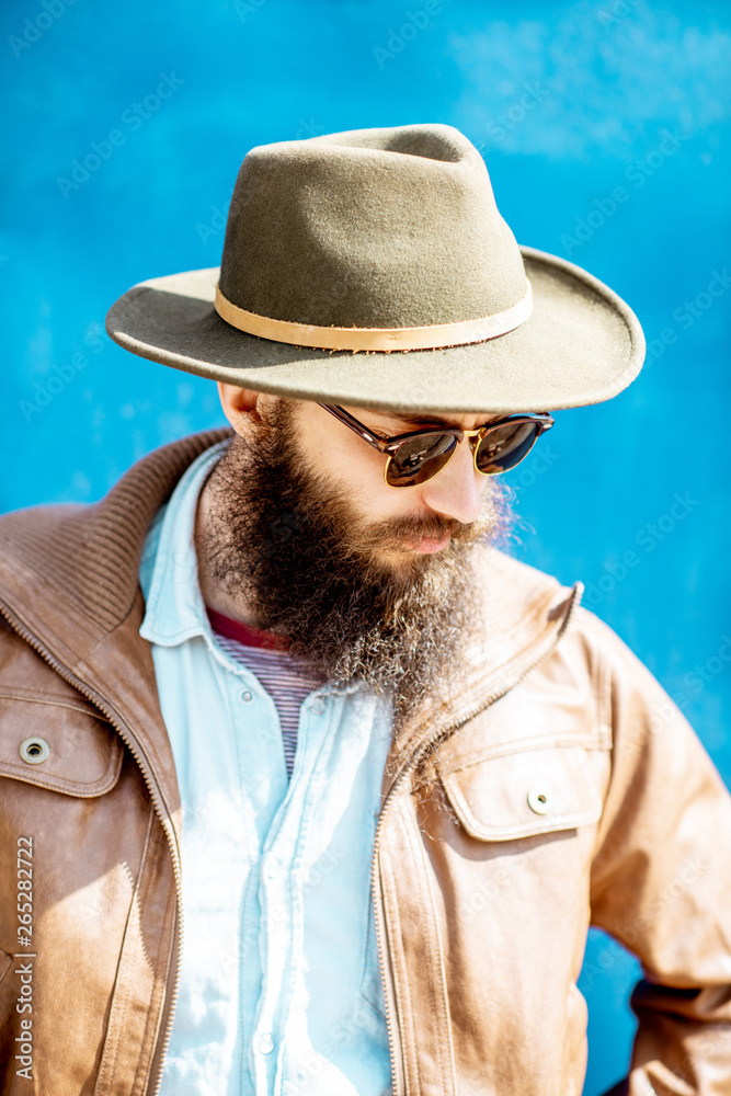 Close-up portrait of a stylish bearded man with hat and sunglasses on the blue wall background