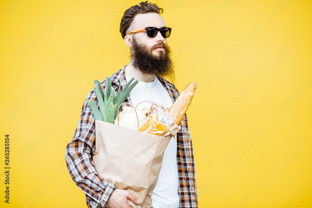 Portrait of a bearded man standing with shopping paper bag full of food on the bright yellow backgro