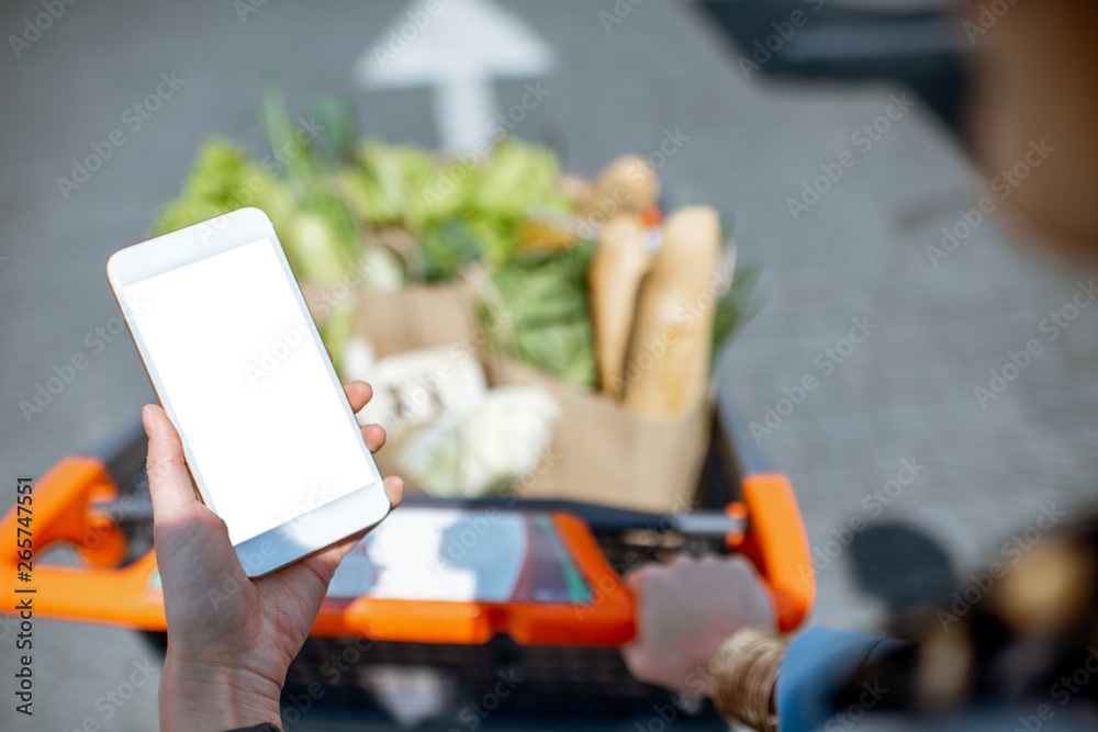 拿着空屏幕的智能手机，背景是装满食物的购物车。网上商店