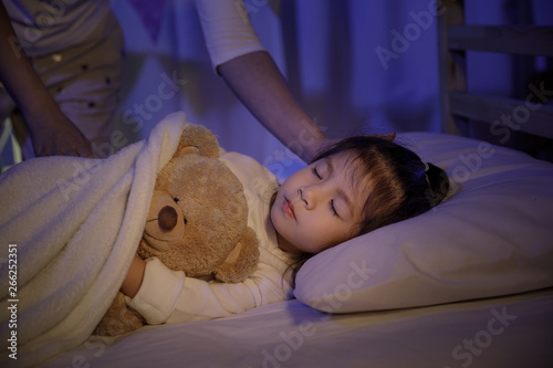 漆黑的卧室里，母亲在床上给小女儿盖上毯子，抚摸着她的头
