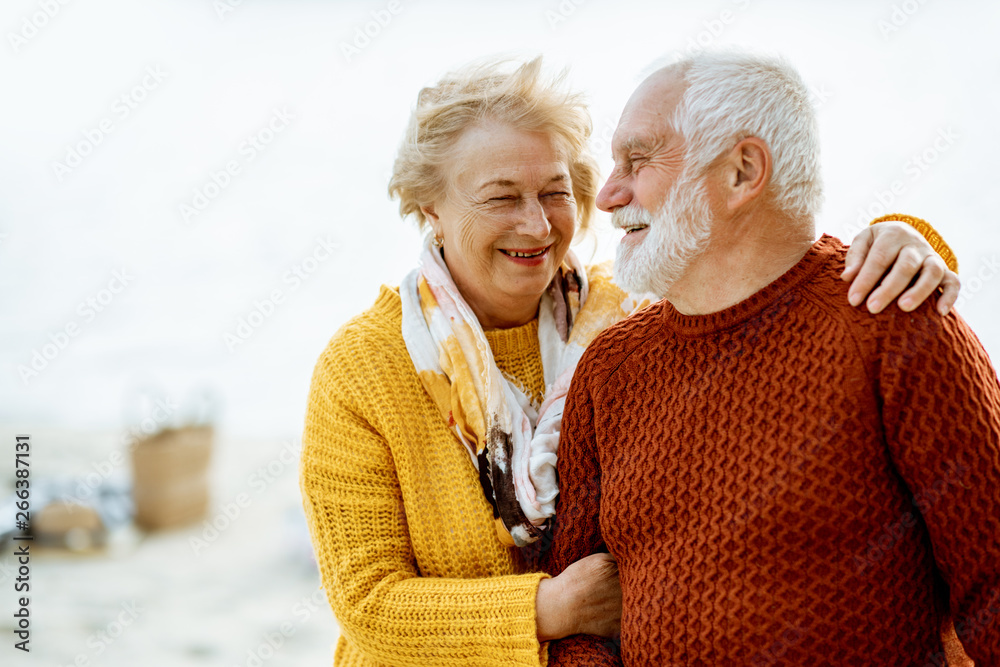 Portrait of a happy senior couple dressed in colorful sweaters hugging on the sandy beach, enjoying 