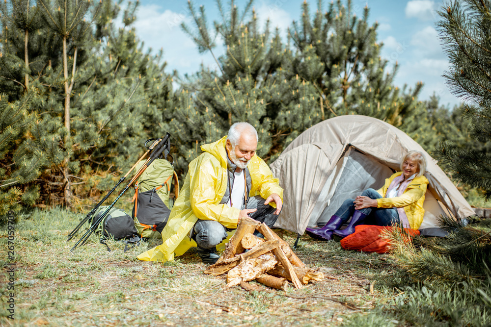 一对穿着黄色雨衣的老年夫妇在树林里帐篷附近的露营地做壁炉