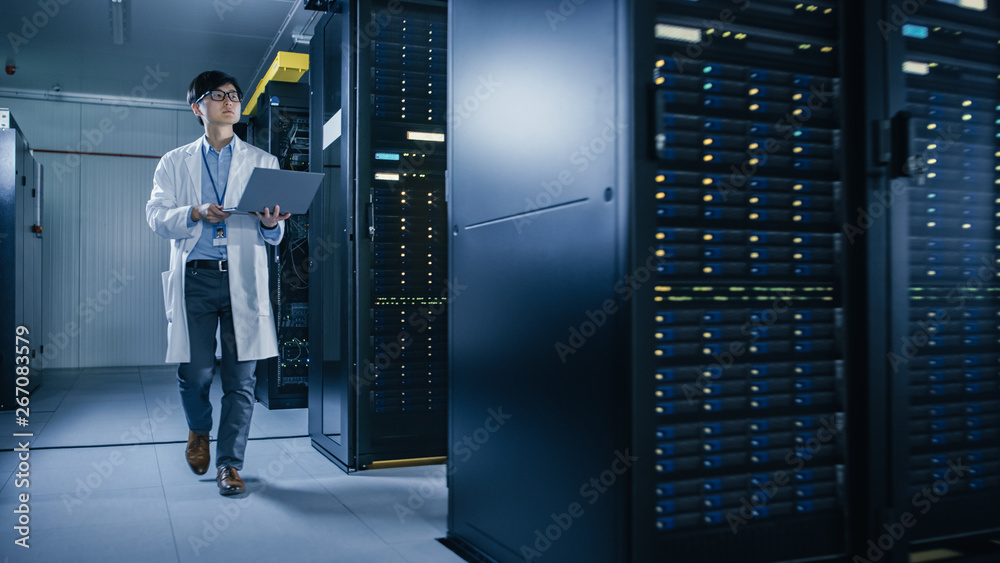 在数据中心：穿着白大褂的男性IT技术人员走在服务器机架旁，使用笔记本电脑公司