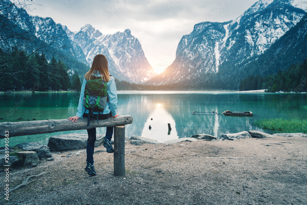 春天日落时，背着背包的年轻女子坐在山湖岸边。在里面旅行