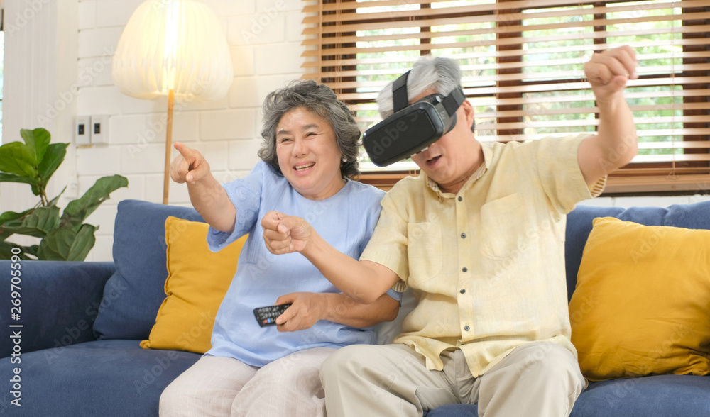 Senior asian couple playing virtual reality headset and using digital tablet in home living room wit