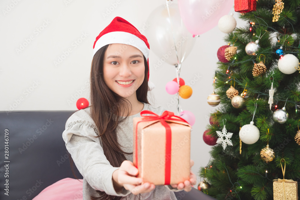 美丽的亚洲女孩戴着圣诞老人帽，在圣诞装饰中微笑着拿着礼盒