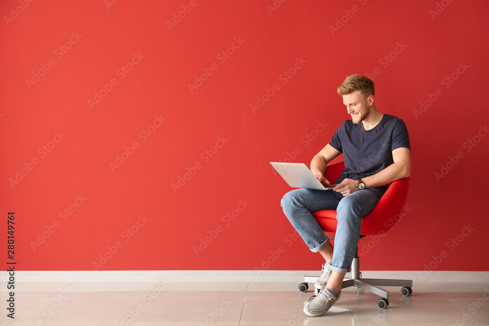 英俊的年轻人拿着笔记本电脑坐在靠近彩色墙的椅子上