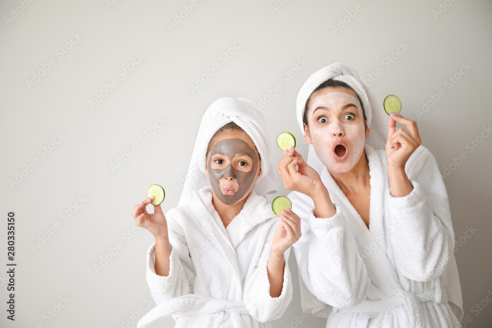 Funny mother and her little daughter with facial masks and cucumber slices on light background