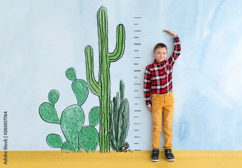 Cute little boy measuring height near color wall with drawn cacti