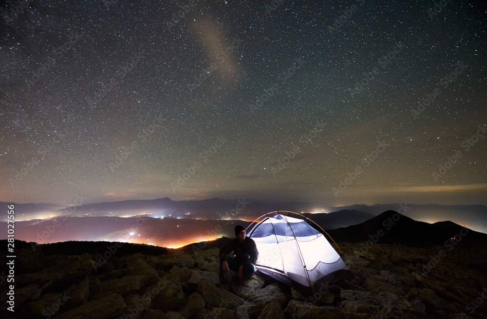 Tourist summer camping at night on the top of rocky mountain. Silhouette of male hiker sitting besid