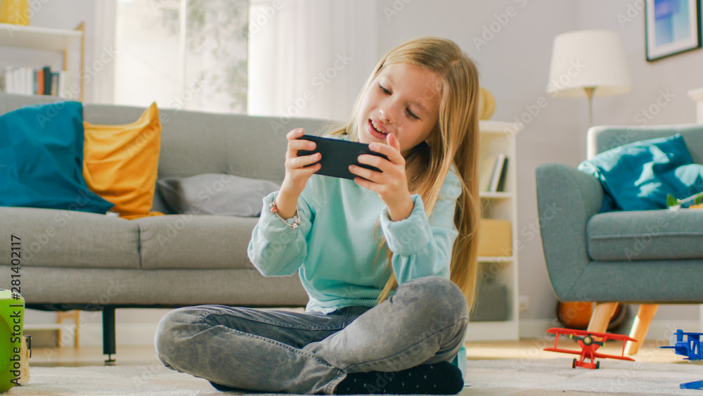 Smart Cute Girl Sitting on a Carpet at Home Playing in Video Game on His Smartphone, Holds and Uses 