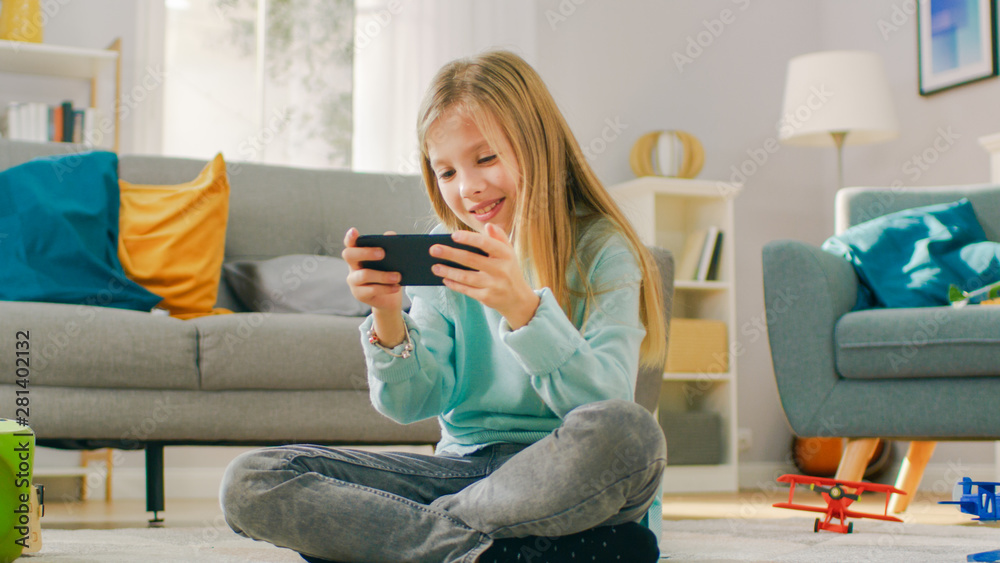 Smart Cute Girl Sitting on a Carpet at Home Playing in Video Game on His Smartphone, Holds and Uses 
