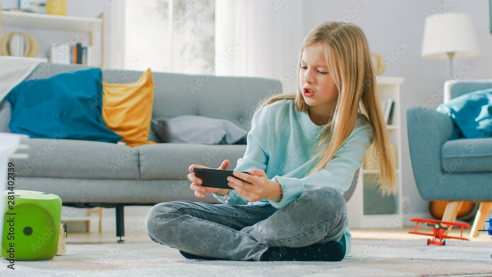 聪明可爱的女孩坐在家里的地毯上用智能手机玩电子游戏，握持并使用