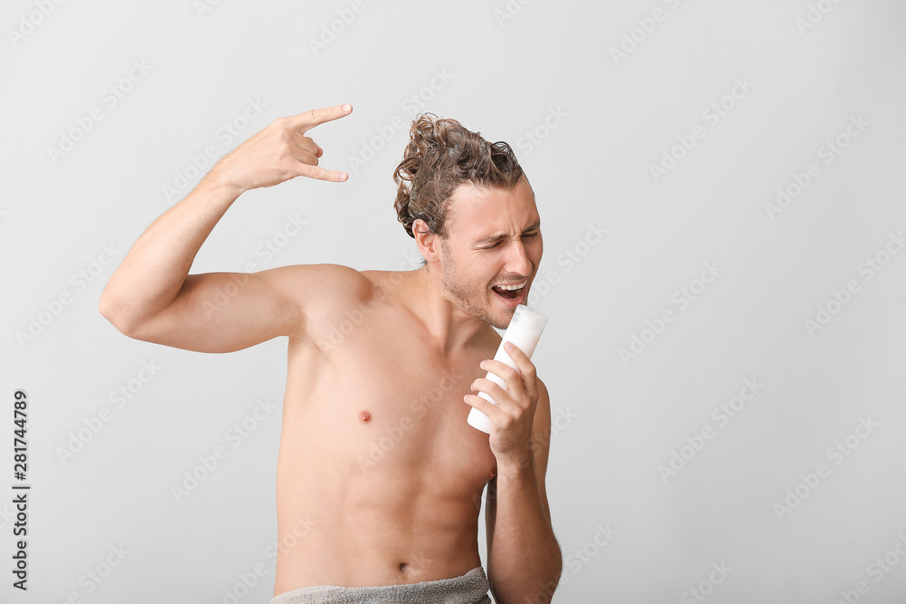 Handsome young man using bottle of shampoo as microphone against grey background