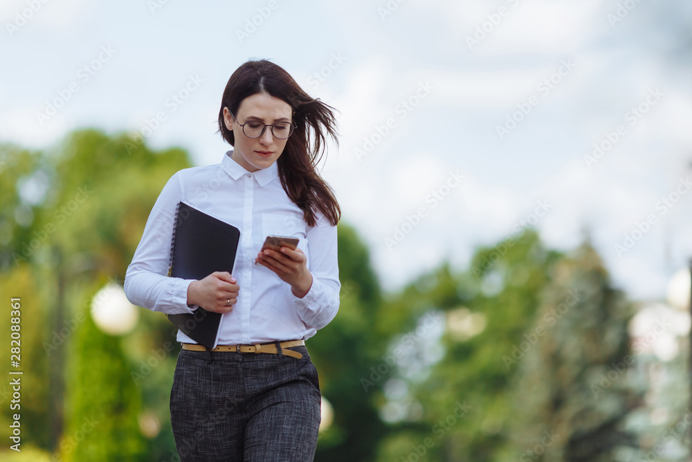 穿着白衬衫的商务女性在城市街道上行走并使用智能手机的正视图