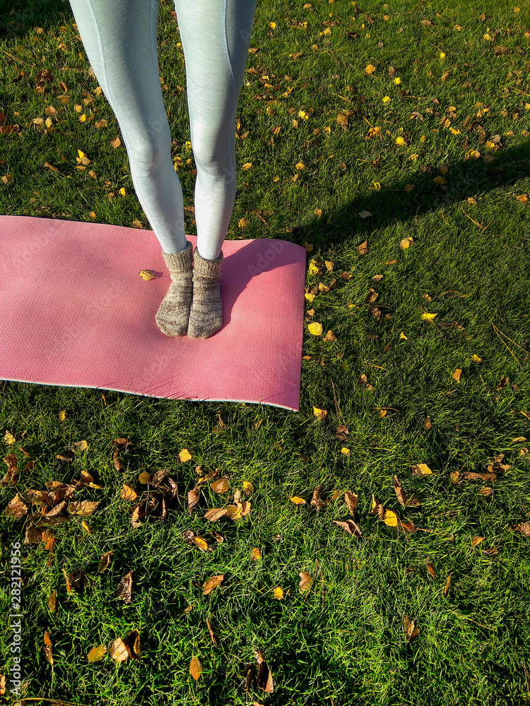 秋天公园里的瑜伽女孩，在黄色落叶的绿色地毯上放着瑜伽垫。有趣的秋天腿