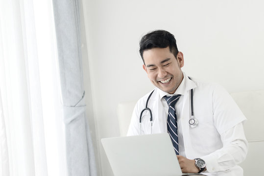Happy Asian doctor using laptop near window, lifestyle concept.