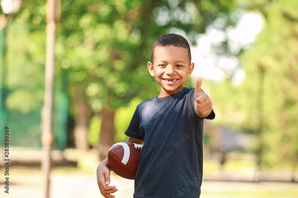 Cute little African-American boy with rugby ball showing thumb-up gesture in park
