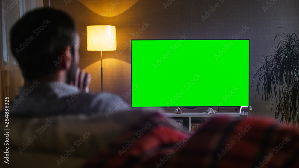 一位中年男子坐在沙发上看大平面电视的晚间背影。