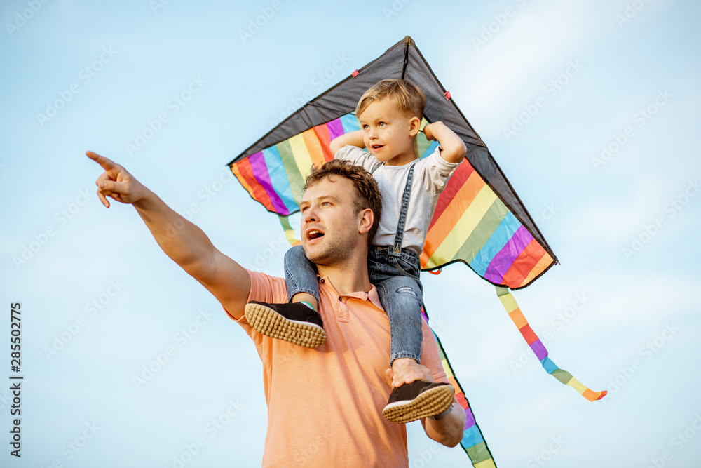 一位幸福的父亲和年幼的儿子的画像，肩上放着五颜六色的空中风筝，在蓝天上