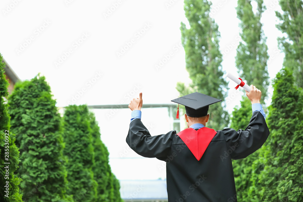 Happy young student in bachelor robe and with diploma outdoors