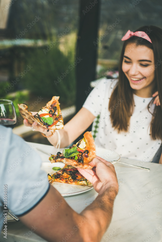 Young happy caucasian couple sitting at table eating freshly baked pizza and drinking wine in Italia