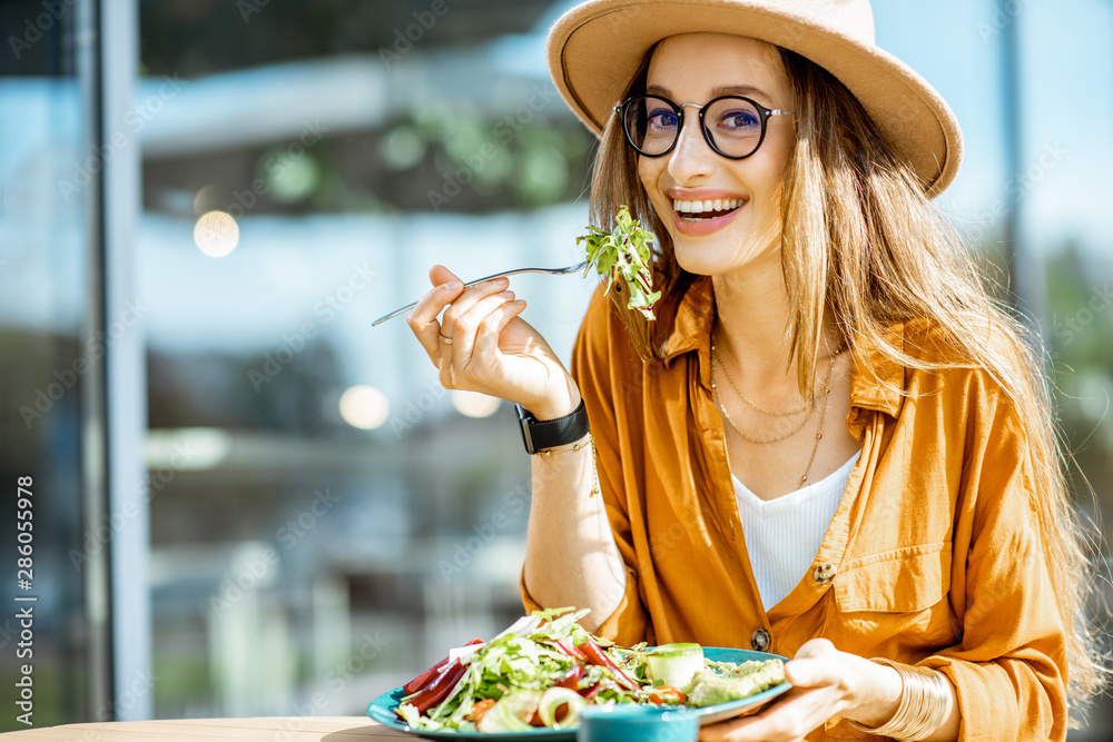 时尚的年轻女性在餐厅露台上吃健康沙拉，在夏日里感到快乐