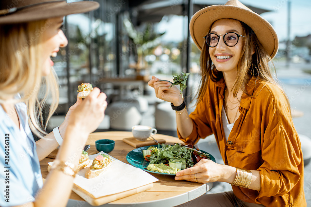 Two female best friends eating healthy food while sitting together on a restaurant terrace on a summ