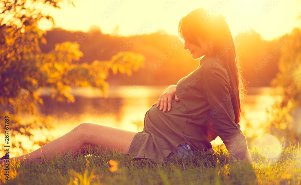 孕妇坐在绿草上，日落时望着河，抚摸着她的肚子。享受治愈