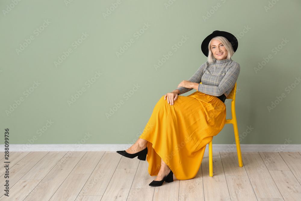 Stylish mature woman sitting on chair against color wall