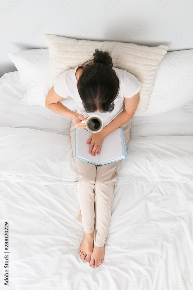 Beautiful young woman drinking coffee while reading book in bedroom, top view