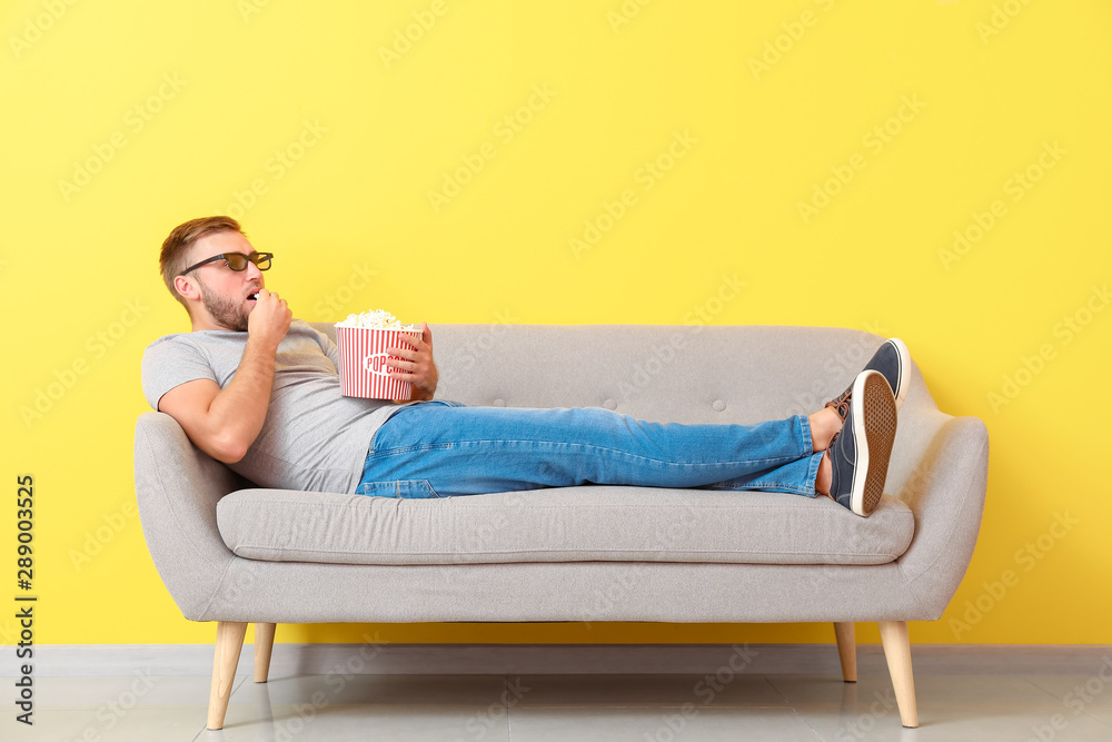 Young man with popcorn watching movie on sofa near color wall