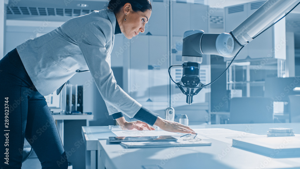 时尚的女性机器人工程师靠在桌子上处理蓝图、文档和平板电脑