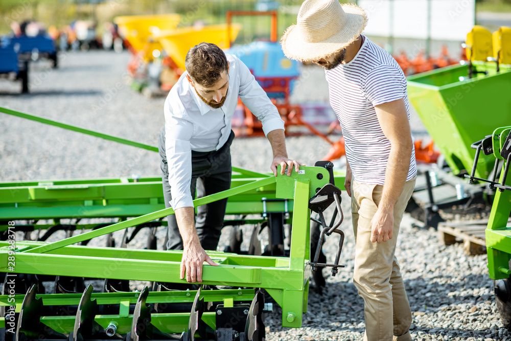 Young agronomist with salesman or manager at the open ground of the shop with agricultural machinery