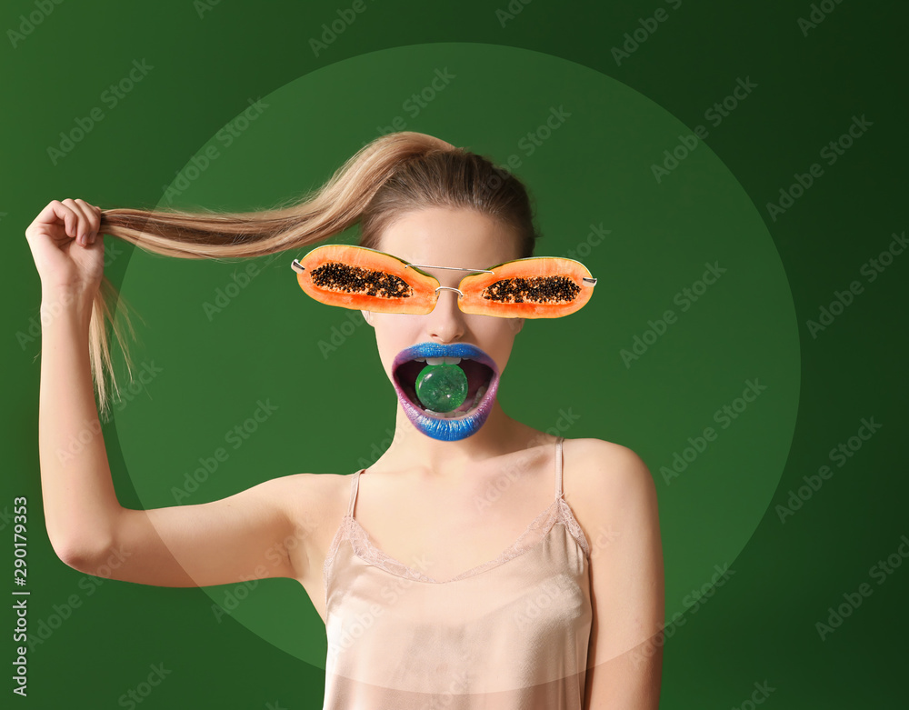 Fashionable young woman with papaya instead her eyes  and candy in mouth on color background